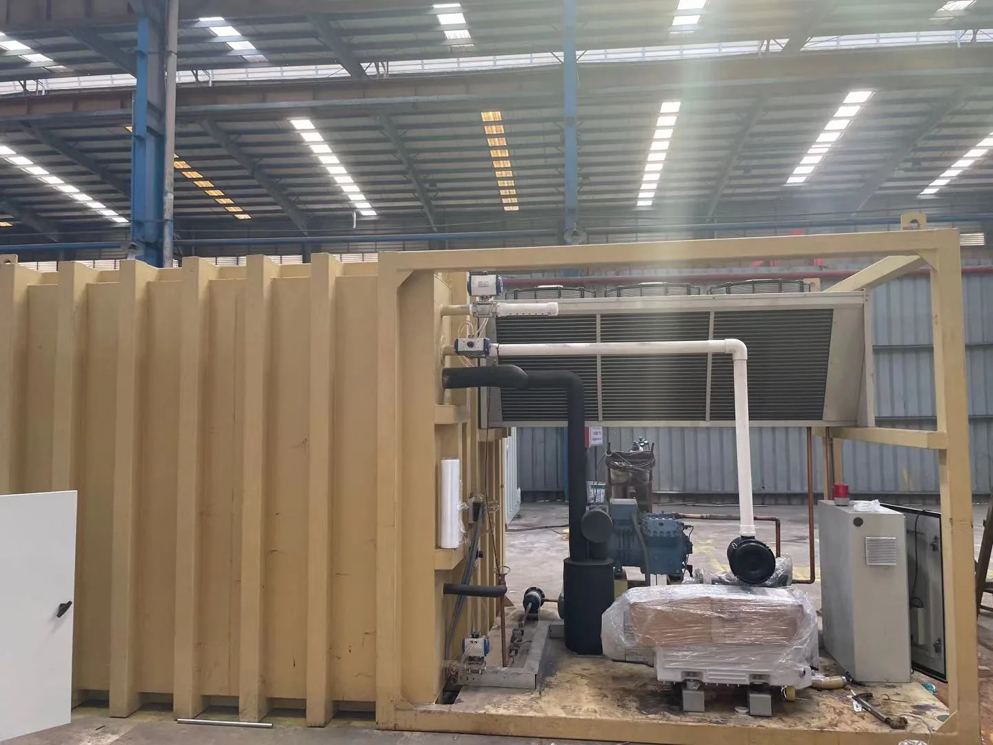 Yellow vacuum chamber beside an open equipment frame with heat exchanger, piping and machinery in a factory