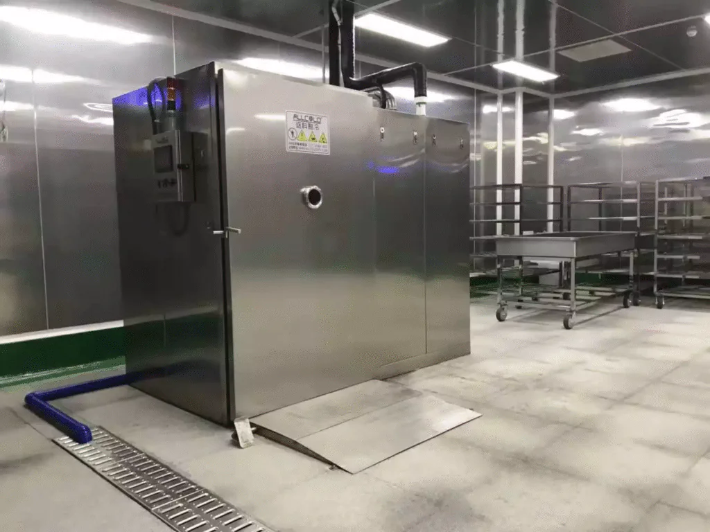 Stainless steel vacuum cooling machine installed in a commercial bakery's clean room, with bakery trays and stainless trolleys in the background.