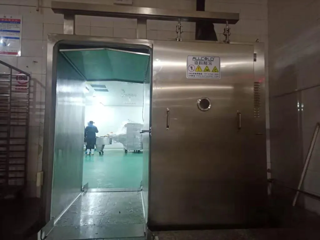 Stainless steel vacuum cooling machine with open door, located in a commercial food facility, showing view into a clean production area with staff.