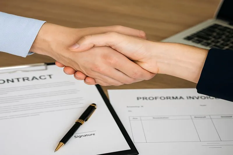 A close-up shot of two hands shaking over a desk with a signed contract and a Proforma Invoice on it, symbolizing a secure deal.