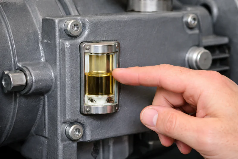 A close-up photo of a finger pointing to the oil sight glass on a vacuum pump, showing clear, golden oil.